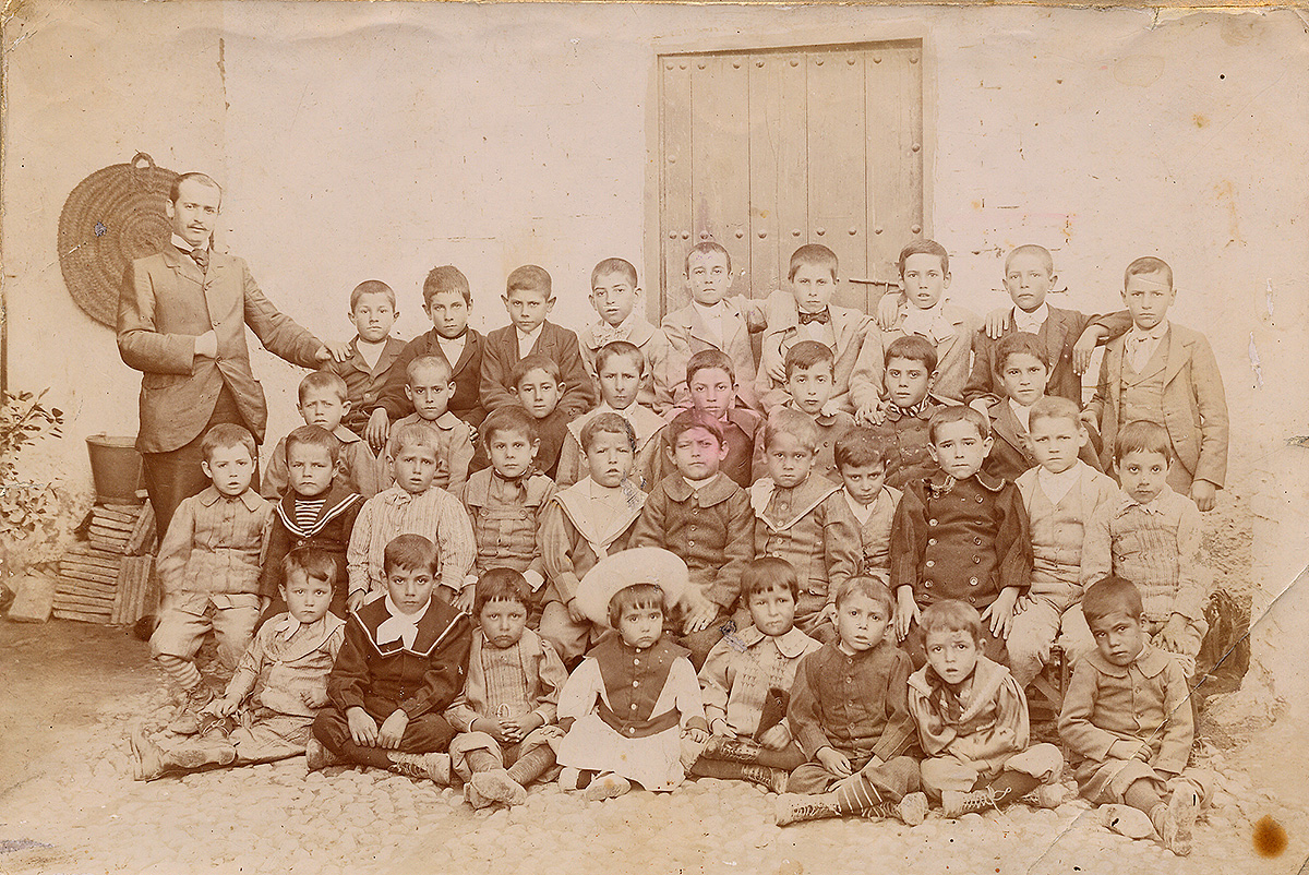 Photo of the students of the kindergarten of Fuente Vaqueros, with Federico García Lorca sitting on the floor and wearing a big hat. Their teacher Antonio Rodríguez Espinosa poses with them.