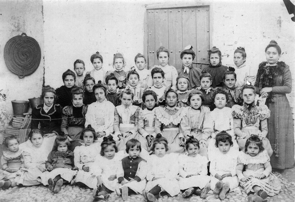 Federico, vestido con chaleco oscuro y sentado en el suelo en el centro, posa con las niñas de la escuela de Fuente Vaqueros y su maestra.
